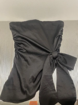 Svart axelbandslös tubtopp med knyt - Snygg svart axelbandslös tubtopp i satinliknande material med stor knytrosett framtill. Toppen har en elegant och stilren look, perfekt för att lyfta vilken outfit som helst. Materialet ger ett lyxigt intryck och toppen sitter tight runt överkroppen.