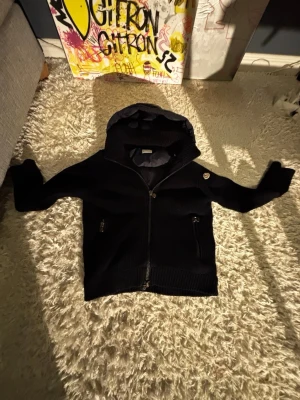 Moncler Cardigan  - Säljer en svart cardigan från Moncler med dragkedja framtill och fickor med dragkedja. Jackan har en ribbad struktur och en klassisk Moncler-logga på ärmen. Perfekt för dig som vill ha en stilren och bekväm jacka med exklusiv känsla.