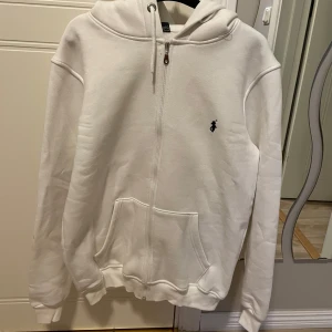 Vit hoodie från Ralph Lauren - Säljer en fräsch vit hoodie från Ralph Lauren med dragkedja och klassisk logga på bröstet. Tröjan har huva, fickor framtill och är tillverkad i mjuk bomull. Perfekt för chill dagar eller när du vill ha en clean look. Storleken är egentligen XL men den sitter som en L!