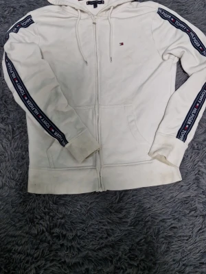 Hoodie Tommy Hilfiger - Vit hoodie med dragkedja från Tommy Hilfiger. Har huva med dragsko, två fickor fram och blå/röda logoband längs ärmarna där det står 'Tommy Hilfiger'. Liten broderad logga på bröstet. Tillverkad i mjuk bomullsmix, perfekt för en avslappnad stil.