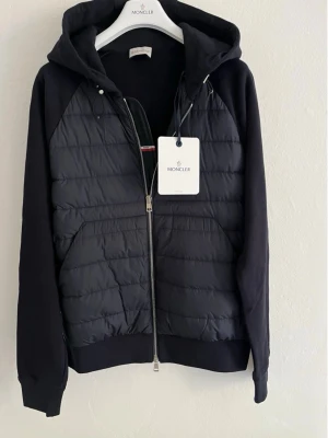 Svart Moncler jacka med huva - Svart jacka från Moncler med quiltad front, huva och dragkedja. Ärmar och rygg i mjuk bomull, medan framsidan är i glansig polyester. Jackan har två fickor framtill och en sportig, stilren look. Perfekt för dig som gillar exklusiva streetwear-vibbar.