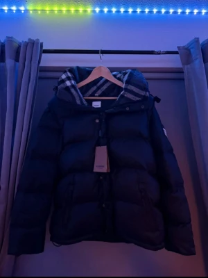 Svart dunjacka med huva från Burberry - Snygg svart dunjacka från Burberry med klassiskt rutigt foder i huvan. Jackan har dragkedja och knappar framtill samt två fickor med dragkedja. Perfekt för kalla dagar och har en modern, puffig look.