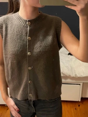 Brun stickad väst med knappar - Mysig brun stickad väst med ribbad halsringning och sex dekorativa knappar framtill. Västen är ärmlös och har en rak, avslappnad passform som passar perfekt över en skjorta eller t-shirt. Materialet ser ut att vara mjukt och lite fluffigt, vilket ger en skön känsla.