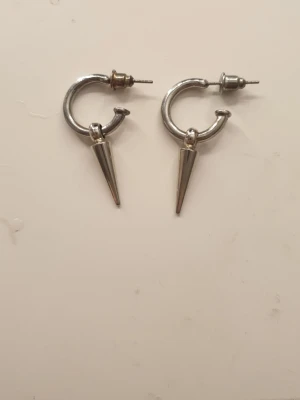 Silvriga spik örhängen - Säljer ett par silvriga örhängen i form av spikar med en cool konformad hängande detalj. Örhängena har en metallisk finish och ger en edgy vibe till din stil. Perfekta för dig som gillar att sticka ut med accessoarer.
