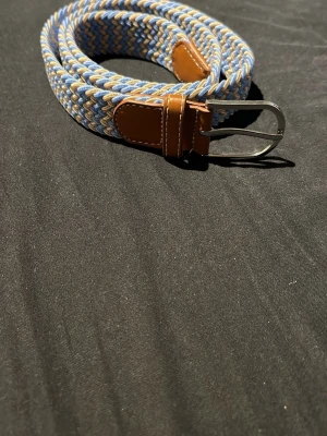 Flätat bälte i blått och beige - Snyggt flätat bälte i blått, beige med detaljer i brunt läder och ett silvrigt spänne. Perfekt accessoar för att lyfta din outfit med en färgklick och modern känsla. Bältet är elastiskt och passar de flesta midjor. 105cm jag har knappast använt bältet.