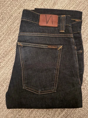 W34/L36 Mörkblå jeans från Nudie Jeans - Snygga mörkblå jeans från Nudie Jeans med klassisk femficksmodell och kontrastsömmar. Jeansen har en slim passform och är tillverkade i slitstarkt bomullsdenim. Perfekta för dig som gillar stilrena och tidlösa jeans. Knappt något märkbart slitage överhuvudtaget. 