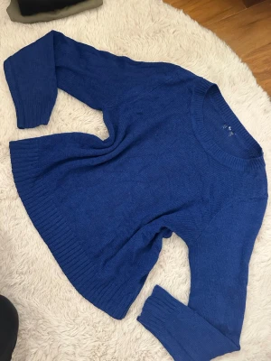 Kornblå stickad tröja med rund hals - En kornblå stickad tröja med rund halsringning och långa ärmar. Tröjan har ribbstickade muddar vid ärmslut och nederkant, vilket ger en snygg och avslappnad look. Obs den har hål vid halsrigning