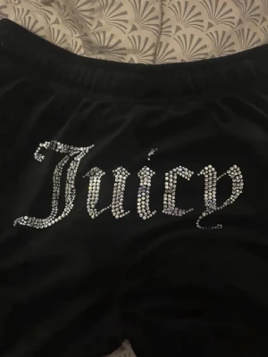 Juicy Couture svart velourset - Säljer ett ikoniskt svart velourset från Juicy Couture med glittrande strasstenar som stavar 'Juicy' på både byxorna och hoodien. Hoodien har dragsko i huvan och muddar vid ärmslut och midja. Perfekt för en chill och trendig look hemma.
