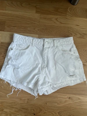 Vita jeansshorts med fransig kant - Säljer ett par vita jeansshorts med råa, fransiga benslut och slitna detaljer framtill. Shortsen har klassisk femficksdesign och knappgylf. Perfekta för sommaren och ger en avslappnad vibe. De har en fläck i höger backficka som är bläck som tyvär inte går bort i tvätten. Har inte testat att få bort de med någon typ av sprej eller liknande. Köpta i Italien märket heter kiabi. Kan sänka priset!!