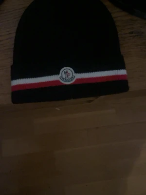 Svart Moncler mössa med ränder - Svart mössa från Moncler med klassisk vit och röd rand längs kanten samt Moncler-logga framtill. Snygg och stilren modell som passar perfekt till kalla dagar. Materialet är stickat och ger en mjuk känsla.