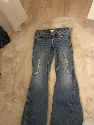 Blå bootcut jeans med broderi - Säljer ett par blå bootcut jeans med slitningar framtill och snygga broderade detaljer på bakfickorna samt längs sidorna. Jeansen har låg midja och klassisk femficksdesign. Perfekta för dig som vill ha en unik och trendig look.