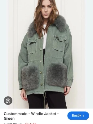 Grön pälsjacka från Custommade - Trendig grön pälsjacka från Custommade med stora grå pälskragar och pälsdetaljer på fickorna. Jackan har knappar framtill, två bröstfickor med lock och dragsko i midjan för en snygg passform. Knappt använd utan defekter