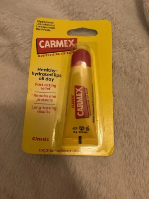 Carmex Classic Läppbalsam tub - Carmex Classic läppbalsam i tub ger intensiv återfuktning och skydd för torra läppar. Tuben är gul med rött lock och röd etikett. Innehåller bland annat kakaosmör, mentol och lanolin för att mjukgöra och reparera läpparna. Perfekt att ha med i väskan.