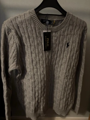 Grå kabelstickad tröja Polo Ralph Lauren - Kabelstickad grå tröja från Polo Ralph Lauren med rund halsringning och klassisk broderad logga på bröstet. Tröjan har långa ärmar och ribbade muddar vid ärmslut och nederkant. Perfekt för dig som gillar stilren och tidlös design.