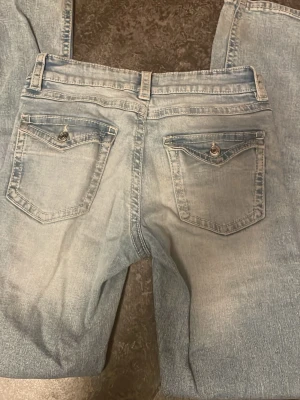 Ljusblå lågmidjade jeans  - Snygga ljusblå jeans med slitningar på knäna och bakfickor med knappdetaljer. Jeansen har en klassisk femficksdesign och är tillverkade i denim. Perfekta för en avslappnad och trendig look.