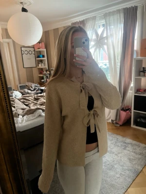 Beige stickad kofta med rosetter - Mysig beige stickad kofta med två knytrosetter framtill. Koftan har lång ärm och rak passform, perfekt att slänga över en topp. Enkel och stilren design som passar till många outfits.