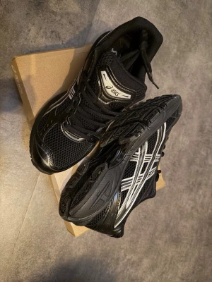 Svarta Asics sneakers med silvriga detaljer - Snygga svarta Asics sneakers med silvriga detaljer och meshpaneler för bra ventilation. Skorna har klassisk snörning och en robust, sportig sula som ger grym komfort. Perfekta för dig som gillar en stilren och sportig look.