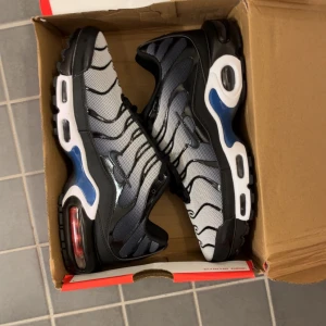 Nike Air Max Plus TN svart/vit/blå - Nike Air Max Plus TN sneakers med svart, vit och blå färg. Ovandel i mesh med svarta vågformade detaljer, synlig Air Max-dämpning i sulan och blåa inslag på sidorna. Klassisk Swoosh-logga och snörning framtill. Perfekta för dig som gillar streetwear och sportig stil.