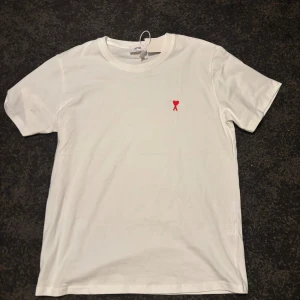 Vit t-shirt från Ami med rött hjärta - Vit t-shirt Ami Alexandre Mattiussi med klassisk passform men skulle säga att den sitter som en L. På bröstet finns ett broderat rött hjärta med bokstaven A. T-shirten är i mjuk bomull och har rund hals och korta ärmar. Perfekt för en clean och stilren look.