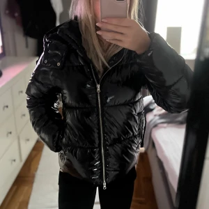 Nelly pufferjacket med huva - Säljer en svart, glansig pufferjacka med huva och dragkedja framtill. Jackan har en quiltad look och är perfekt för kalla dagar. Snygg och trendig modell som håller dig varm och stilren.