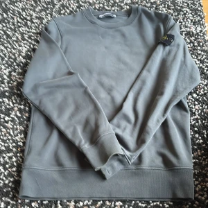 Grå Stone Island sweatshirt, strl S i mkt bra skick  - Säljer en grå sweatshirt från Stone Island med klassisk rund hals och ribbade muddar. Tröjan har den ikoniska Stone Island-patchen på vänster ärm. Perfekt basic med snygg streetkänsla och stilren design. Storleken är S, äkta vara. Tröjan är i mkt bra skick 