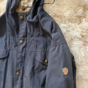 Blå parkajacka från Fjällräven - Snygg blå parkajacka från Fjällräven med klassisk huva, tryckknappar och flera fickor framtill. Jackan har rutigt innerfoder och ikonisk Fjällräven-logga på ärmen. Perfekt för dig som gillar outdoor och streetstyle.