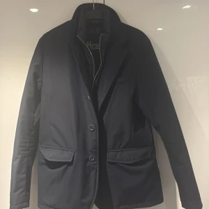 Svart dunfodrad kavajjacka från Herno - Stilren svart kavajjacka från Herno Sub-Zero med dunfoder och vattenavvisande yttertyg. Jackan har klassisk kavajkrage, tre knappar framtill, fickor med lock och quiltad insida med dragkedja. Perfekt för kalla dagar när du vill vara både varm och snygg.