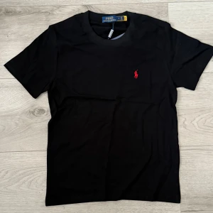 Svart T-shirt med röd logga - Säljer en snygg svart t-shirt med en röd logga vid vänstra bröstet. Helt oanvänd och erbjuder ett prisvärt erbjudande för denna T-shirt, perfekt till dig som vill ta din klädstil till nästa nivå. Frågor, Dma👍🏼😁