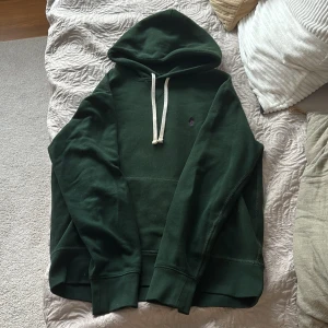 Grön hoodie från Polo Ralph Lauren - Mörkgrön hoodie från Polo Ralph Lauren med lila broderad logga på bröstet. Tröjan har huva med vit snörning och en klassisk känguruficka framtill. Tillverkad i mjuk bomull, perfekt för chill dagar. Passformen är normal och ärmarna är långa.