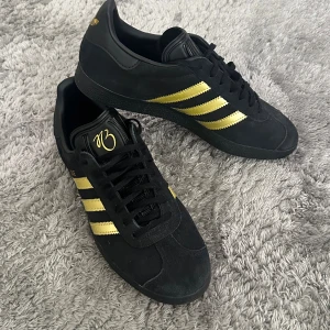 Adidas Gazelle svart/guld sneakers Jude Bellingam - Adidas Gazelle sneakers i svart mocka med guldiga detaljer och klassiska tre ränder på sidan. Snyggt broderad logga på plösen och insidan har en cool grafisk figur. Platt sula och rund tå, snörning framtill. Perfekt för dig som vill sticka ut med stil.