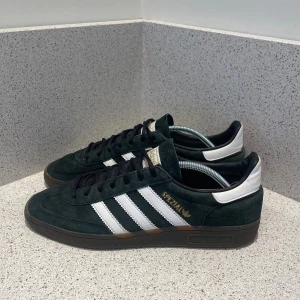 Adidas Spezial svarta sneakers - Adidas Spezial sneakers i mörkgrönsvart mocka med vita läderdetaljer och klassiska tre ränder på sidan. Skorna har svart snörning, bruna gummisulor och rund tå. Guldtexten 'Spezial' på sidan ger en extra touch. Perfekt för dig som gillar retrostil och streetwear.