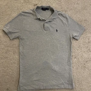 Grå pikétröja från Polo Ralph Lauren - Snygg grå pikétröja från Polo Ralph Lauren i klassisk passform. Tröjan har korta ärmar, krage och knäppning framtill. På bröstet finns den ikoniska broderade loggan i mörkblått. Perfekt för dig som gillar stilrena och tidlösa plagg. Den är i s men passar mer som m