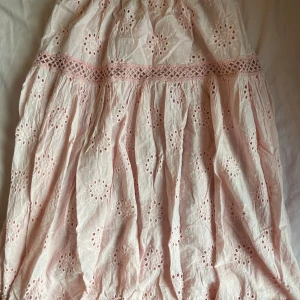 Ljusrosa broderad kjol med spets - Supersöt ljusrosa kjol i bomull med broderade blommor och fina spetsdetaljer. Kjolen har en luftig och lätt känsla, perfekt för varma dagar. Den har vackra hålbroderier och dekorativa band i spets som ger en romantisk vibe.