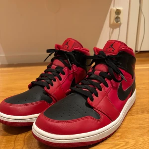 Nike Air Jordan 1 röd/svart sneakers - Säljer ett par Nike Air Jordan 1 sneakers i klassisk röd och svart färg med vita sulor. Skorna har svart snörning, ikonisk Swoosh-logga och Air Jordan Wings-märke på sidan. Tillverkade i skinn med hög mid-cut och platt sula. Perfekta för dig som vill sticka ut med en tidlös streetstyle. Skorna är knappt använda.