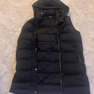 Svart dunväst  - Svart dunväst från Ullared i storlek M (40-42). Västen har en enkel design, dragkedja framtill, fickor med dragkedja och en huva med snörning. Perfekt för höst och vinter! 