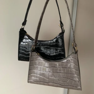 Grå och svart axelväska med krokoprägling - Två stilrena axelväskor i grått och svart med glansig krokopräglad yta och guldfärgade detaljer. Väskorna har en smal, justerbar axelrem och stängs med dragkedja. Perfekt storlek för det viktigaste och ger en cool vibe till din outfit.