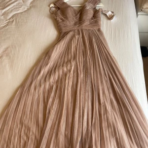 Champagnefärgad långklänning med glitter - Säljer en elegant champagnefärgad långklänning från Pxxi med glittriga detaljer. Klänningen har v-ringad omlottliv, breda axelband och plisserad kjol som ger ett lyxigt flöde. Perfekt för bal eller nyår! Materialet är skimrande syntet med mjuk känsla.