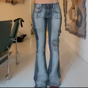 Blekta jeans med fickor - Säljer ett par blå bootcut cargojeans med flera stora fickor och coola detaljer. Jeansen har en tvättad look och utsvängd design. Perfekt för dig som gillar unika jeans. Tyvärr saknas en ficka men det ger egentligen bara jeansen lite mer känsla! 