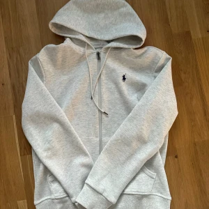 Grå hoodie från Polo Ralph Lauren - Ljusgrå hoodie från Polo Ralph Lauren med dragkedja framtill, huva med snörning och klassisk broderad logga på bröstet. Tillverkad i mjuk bomullsblandning och har fickor på sidorna. Perfekt för en chill och stilren look.