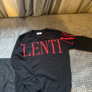 Svart stickad tröja Valentino - Svart stickad tröja från Valentino med stora röda bokstäver över bröstet och på båda ärmarna. Rund halsringning och långa ärmar med ribbade muddar. Tröjan har en clean look med en tydlig logga som sticker ut. Perfekt för dig som gillar statement-plagg.