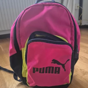 Färgglad ryggsäck från Puma - Cool ryggsäck från Puma i stark rosa, gul och svart. Perfekt för skolan eller häng med kompisar. Puma-logga framtill och bekväma vadderade remmar. Liten model