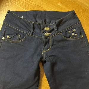 Mörkblå skinny jeans med snörning - Snygga mörkblå skinny jeans från Miss Sixty, ventury med gul kontrastsöm, dubbla knappar och coola detaljer som snörning och nitar bak. Jeansen har låg midja och smal passform, perfekt för dig som gillar att sticka ut med din stil.