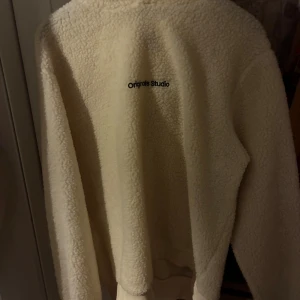 Beige teddyjacka från Originals Studio - Mysig beige teddyjacka från Originals Studio med broderad logga på ryggen. Jackan har lång ärm och är tillverkad i fluffigt fleece-material som håller dig varm under kyliga dagar. Perfekt för dig som gillar en chill och trendig stil.