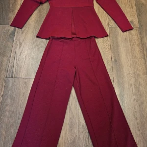 Vinröd kostym med puffärm och vida byxor - Snygg vinröd kostym med kavaj som har puffärmar, v-ringning och slits framtill. Byxorna är vida och har pressveck för en snygg look. Passar mycket bra och ser eleganta ut. Byxorna har lite lossning upptill (se bild) men syns inte och kan enkelt sys.