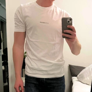 Vit t-shirt från C.P. Company - Vit t-shirt från C.P. Company med svart logga tryckt på bröstet. Klassisk rund halsringning och normal passform. Tillverkad i mjuk bomull som känns skön mot huden. Perfekt för en clean och stilren look. Använd 2-3 gånger. 