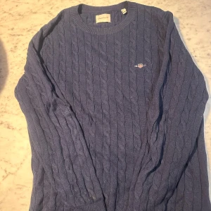 Blå kabelstickad tröja från GANT - Säljer en klassisk blå kabelstickad tröja från GANT med rund halsringning och broderad logga på bröstet. Tröjan har långa ärmar och ribbade muddar, perfekt för lager-på-lager under kyliga dagar.
