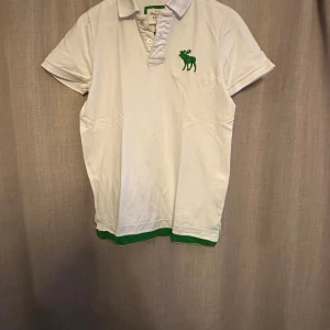 Vit pikétröja Abercrombie & Fitch  - Vit pikétröja från Abercrombie & Fitch med grön logga på bröstet och grön detalj längst ner. Klassisk krage och korta ärmar, perfekt för en clean och sportig look. Tillverkad i mjuk bomull för skön känsla. 