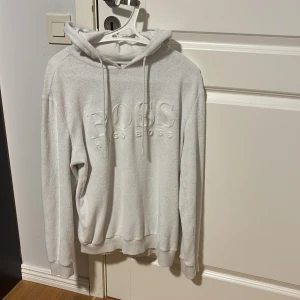 Vit hoodie från Hugo Boss - Säljer en stilren vit hoodie från Hugo Boss med broderad logga framtill. Tröjan har huva med snörning, långa ärmar och ribbade muddar. Perfekt för dig som gillar en clean och exklusiv look. Storlek S men passar även M