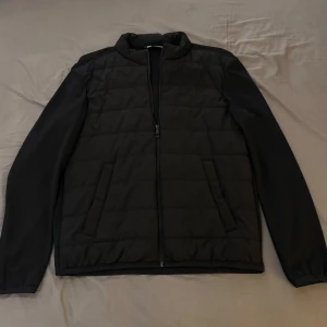 Svart quiltad jacka från Zara Man - Stilren svart jacka från Zara Man med quiltad front och släta ärmar. Jackan har dragkedja framtill, två snedställda fickor och en enkel ståkrage. Perfekt för dig som gillar en clean look och vill ha något lätt men ändå värmande.