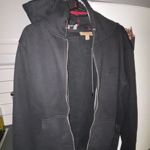 Svart hoodie från Lonsdale med dragkedja - Svart hoodie från burberry  med dragkedja och huva med snörning. Klassisk logga broderad på bröstet och två fickor framtill. Insidan av huvan har ett rutigt mönster i svart, vitt och rött. Perfekt för chill dagar eller när du vill ha en clean streetstyle. Ny skick använd en gång 